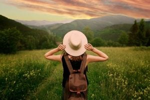 Woman with hat and backpack standing in field looking at view of mountains